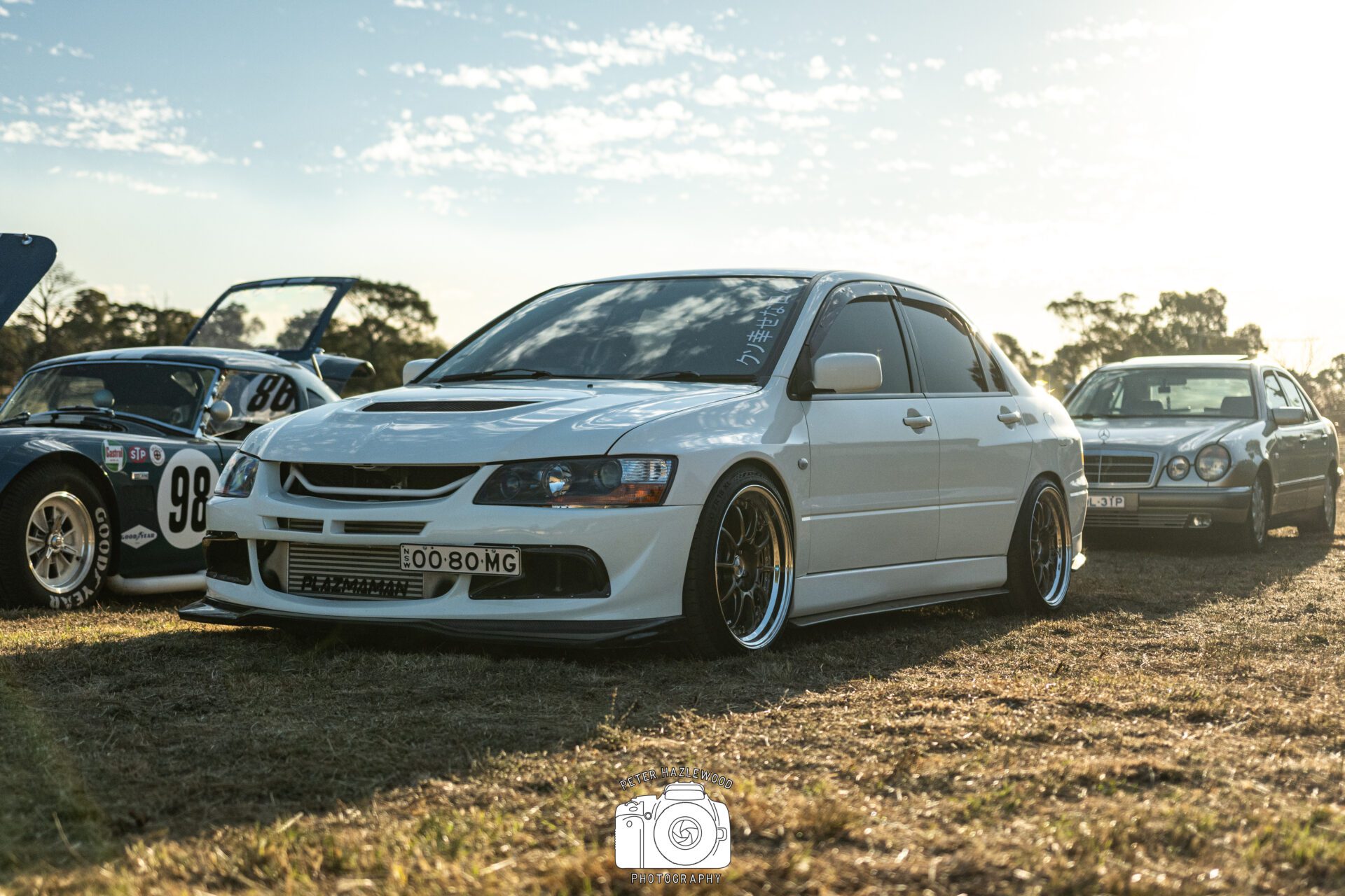 White modified sedan with lowered stance parked on grass, surrounded by classic cars under a clear sky.