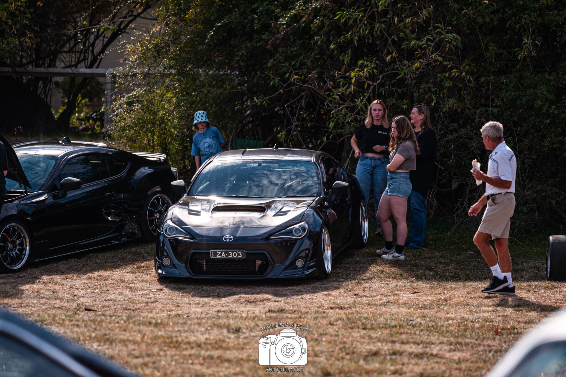 Black Toyota sports car parked on grass with people standing and chatting nearby.