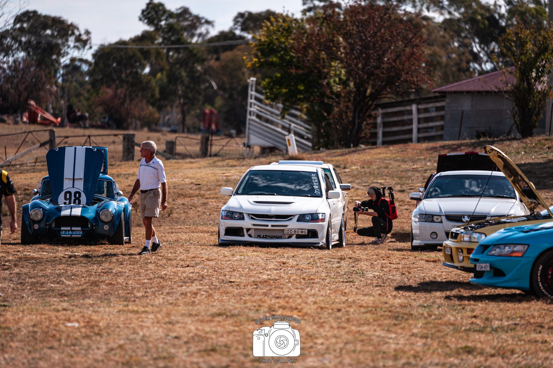 Car show on dry grass with classic and modern cars, some with hoods open and people around.