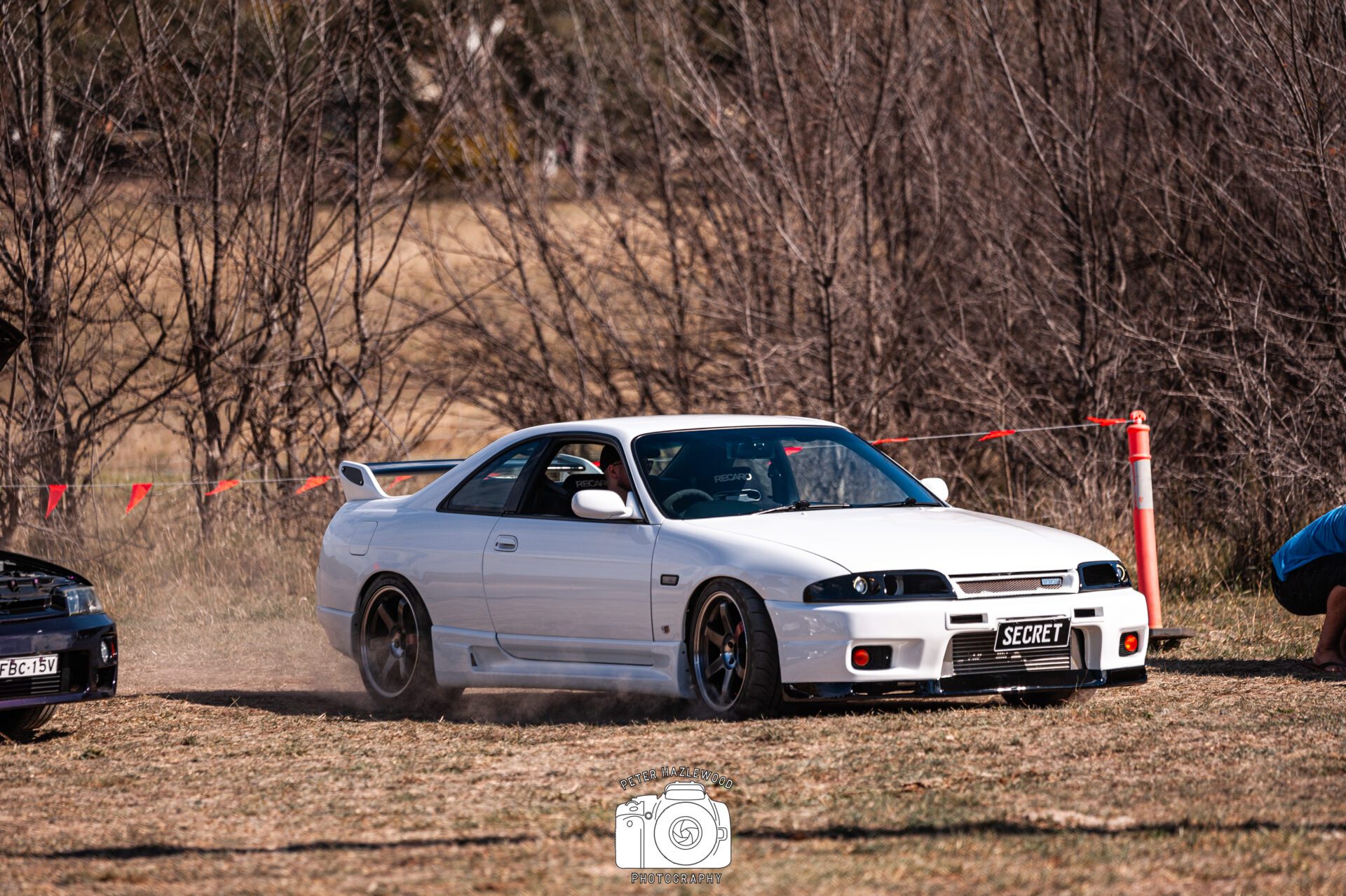 White sports car drifting on a grassy area near bare trees with orange safety cones and tape.