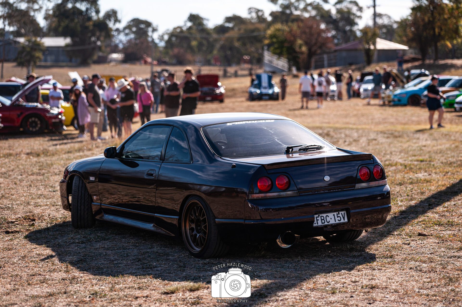 Black Nissan Skyline car parked on grass with people and other cars in background at event.