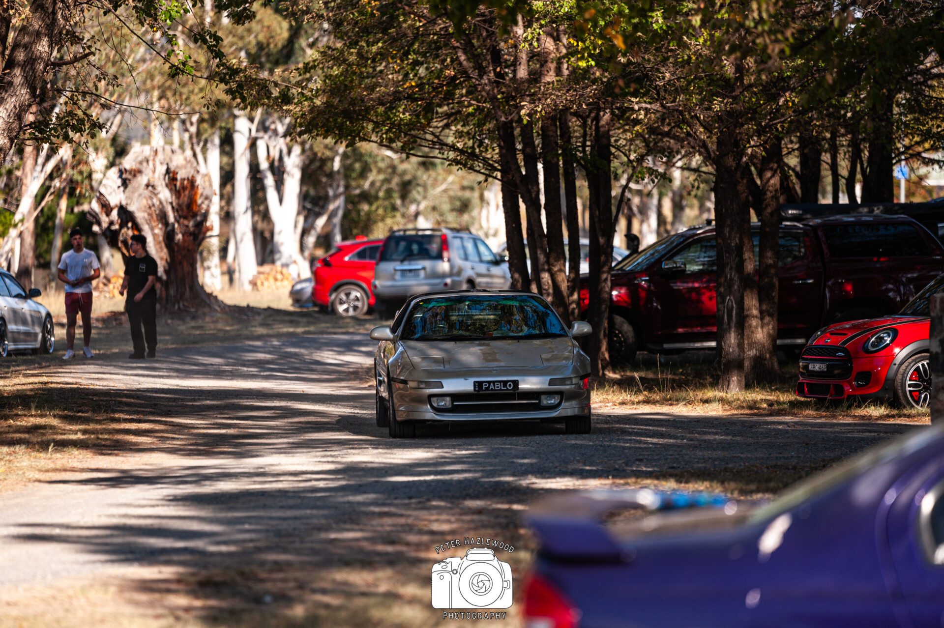 Silver sports car with "PABLO" license plate driving on a tree-lined dirt road, people walking nearby.