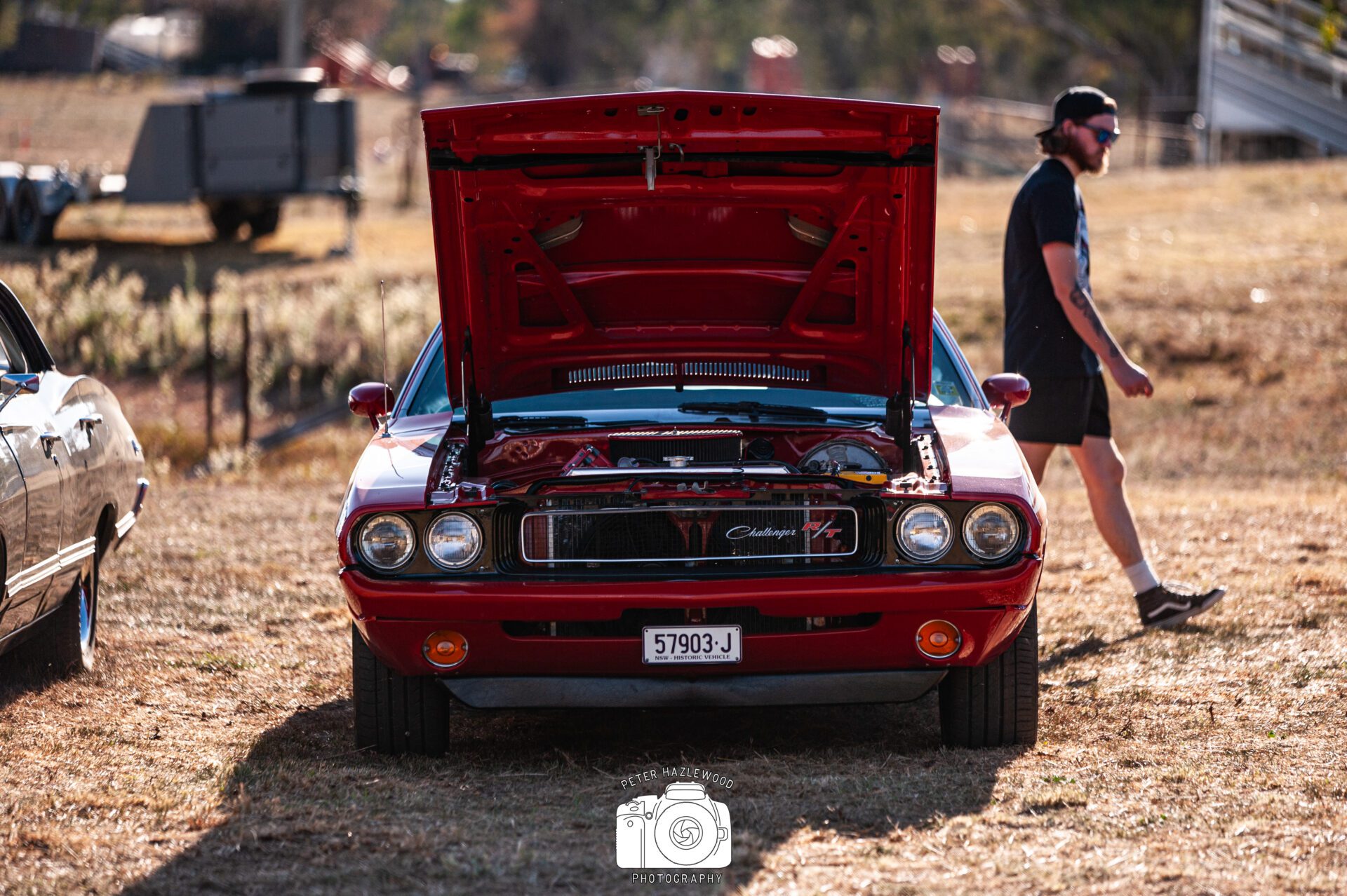 Red Dodge Challenger R/T with hood open parked on dry grass, person walking nearby.