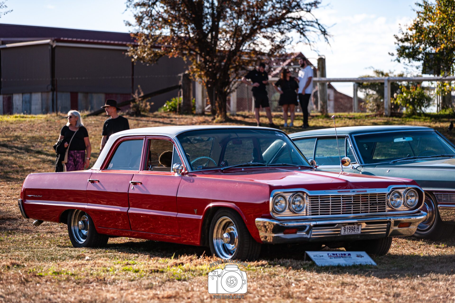 Red and white classic Chevrolet Impala parked on grass at an outdoor car show with people in background.
