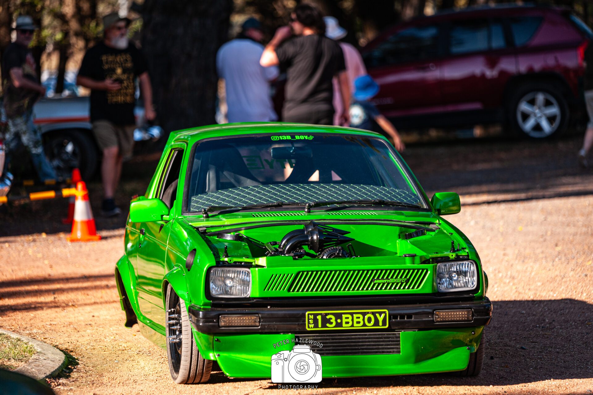 Bright green modified car with exposed engine at an outdoor event, people in background.