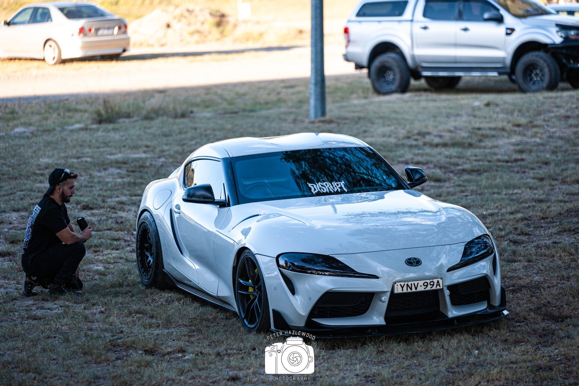 White Toyota Supra parked on grass with a person crouching nearby holding a drink.