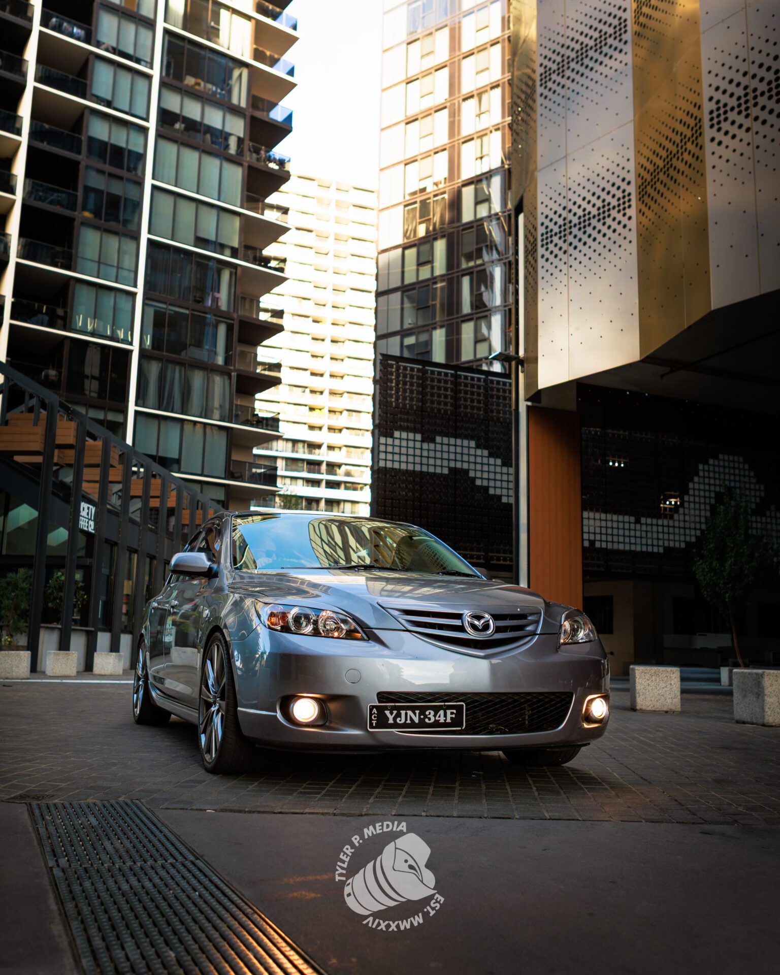 Silver Mazda sedan with headlights on parked in an urban area surrounded by tall buildings.