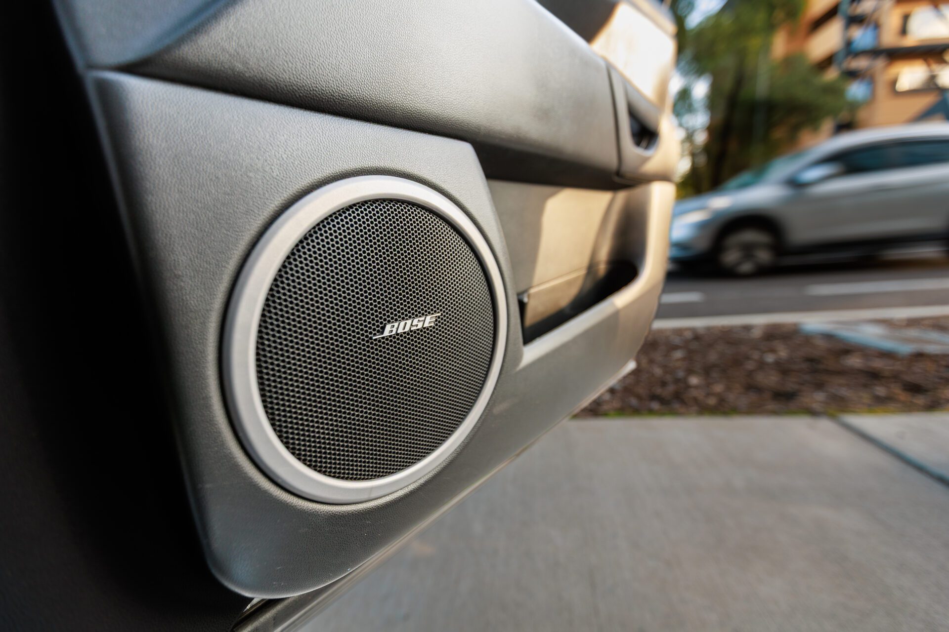 Close-up of a Bose speaker installed in a car door panel with a blurred street background.