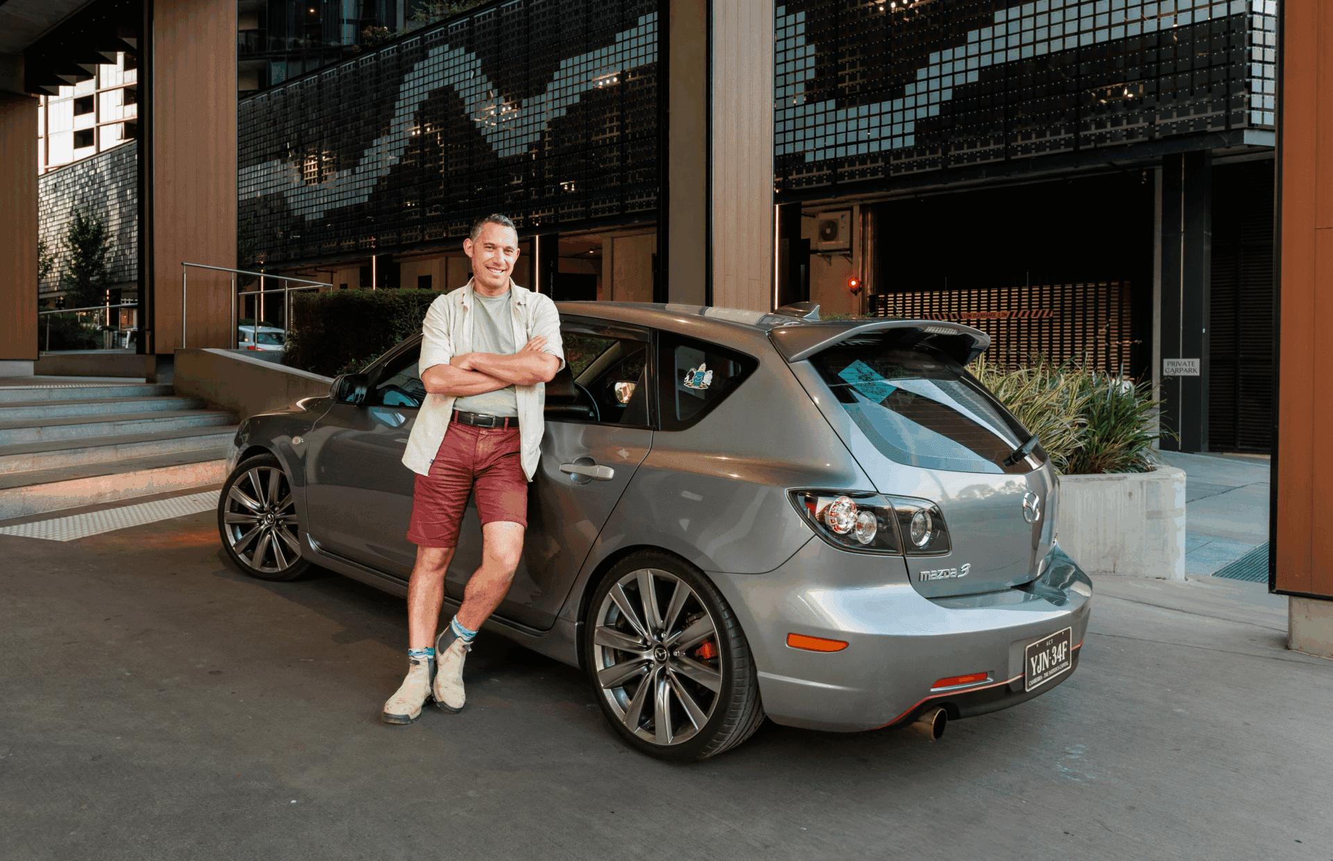 Man in red shorts leans on a silver Mazda 3 hatchback parked in an urban setting.