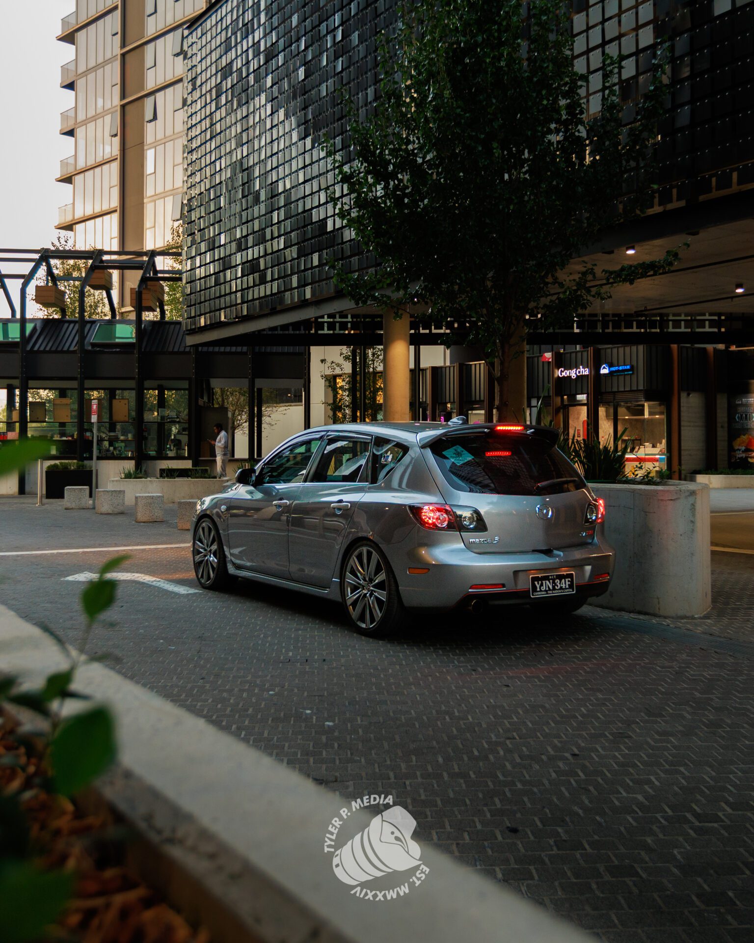 Silver Mazda hatchback parked on a city street near modern buildings at dusk.