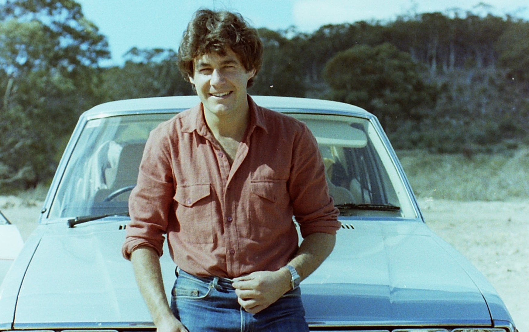 Young man in red shirt leaning on white car outdoors with trees in background.