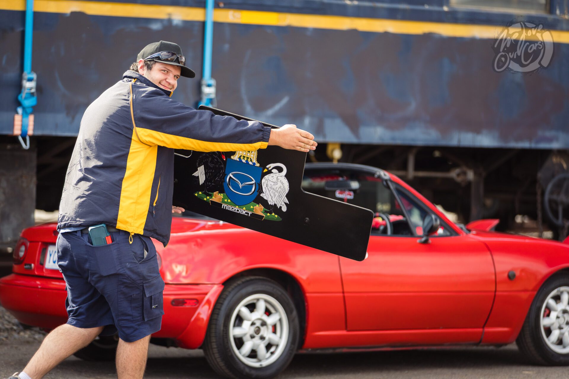 Man holding a car spoiler near a red convertible sports car in an outdoor setting.