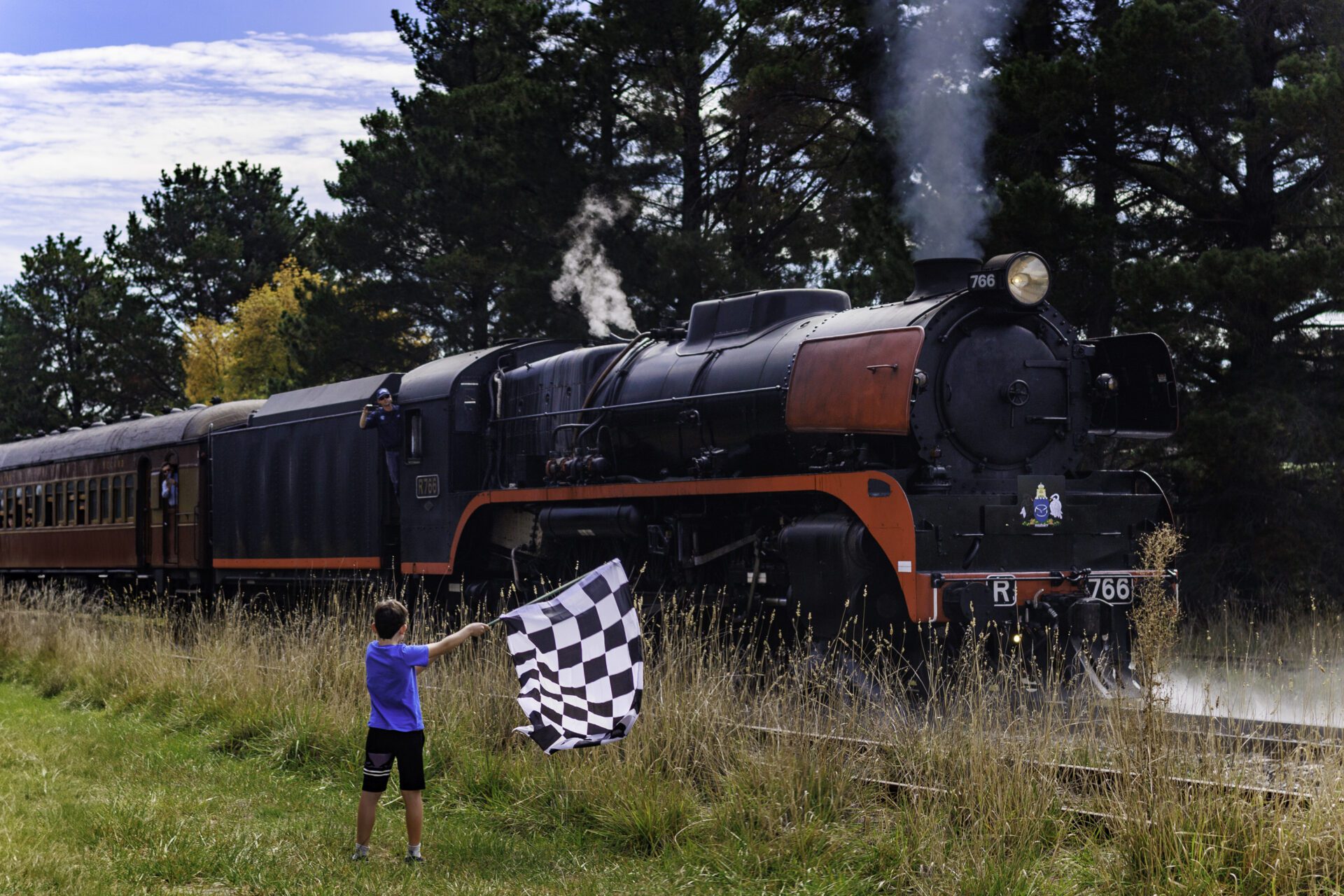 Boy waving checkered flag near a vintage steam train moving through grassy trackside.