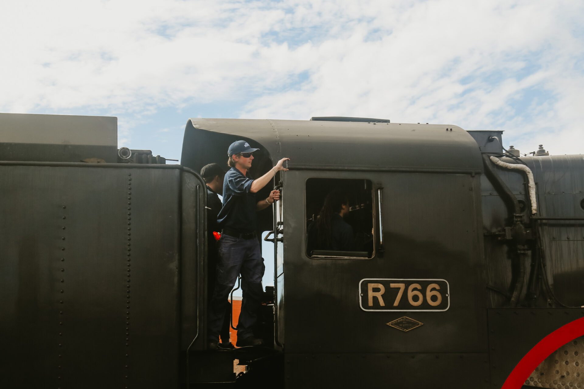 Person standing on the steps of a black steam locomotive marked R766, with others inside the cabin.