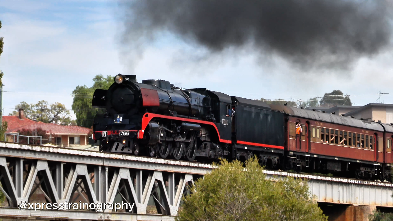 Black steam locomotive with red trim crossing a metal bridge, emitting thick black smoke.