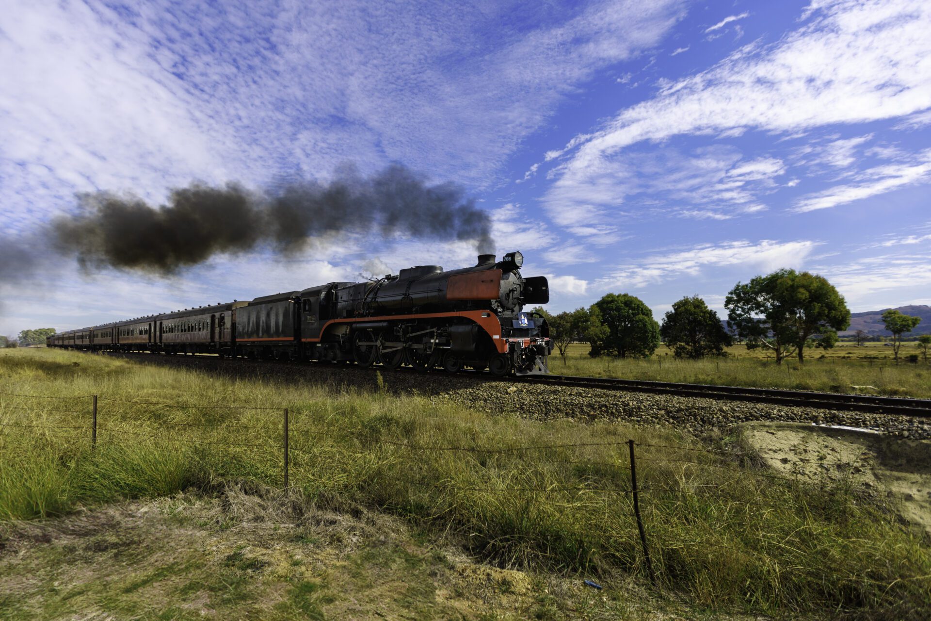Black steam train with red accents emits smoke, moving on tracks through grassy field under blue sky.