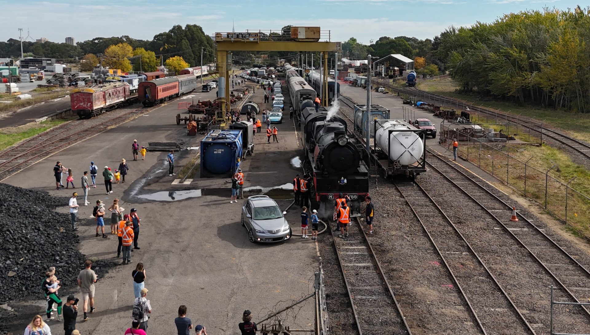 Steam train with people and staff in orange vests at a busy train yard on a partly cloudy day.