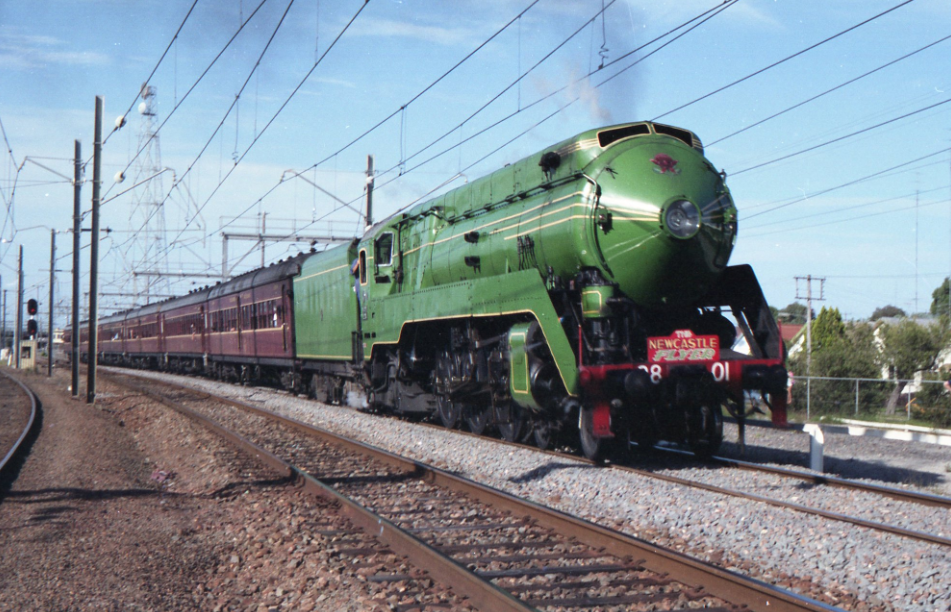 Green vintage steam locomotive pulling maroon passenger cars on railway tracks under blue sky.