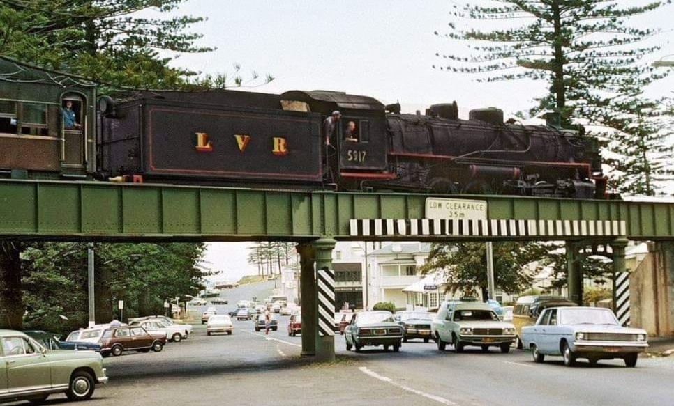 Steam locomotive crossing a green railway bridge over a busy street with vintage cars below.