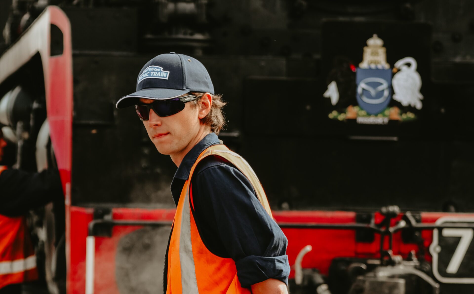 Man wearing sunglasses, cap, and orange safety vest standing in front of a black and red train.