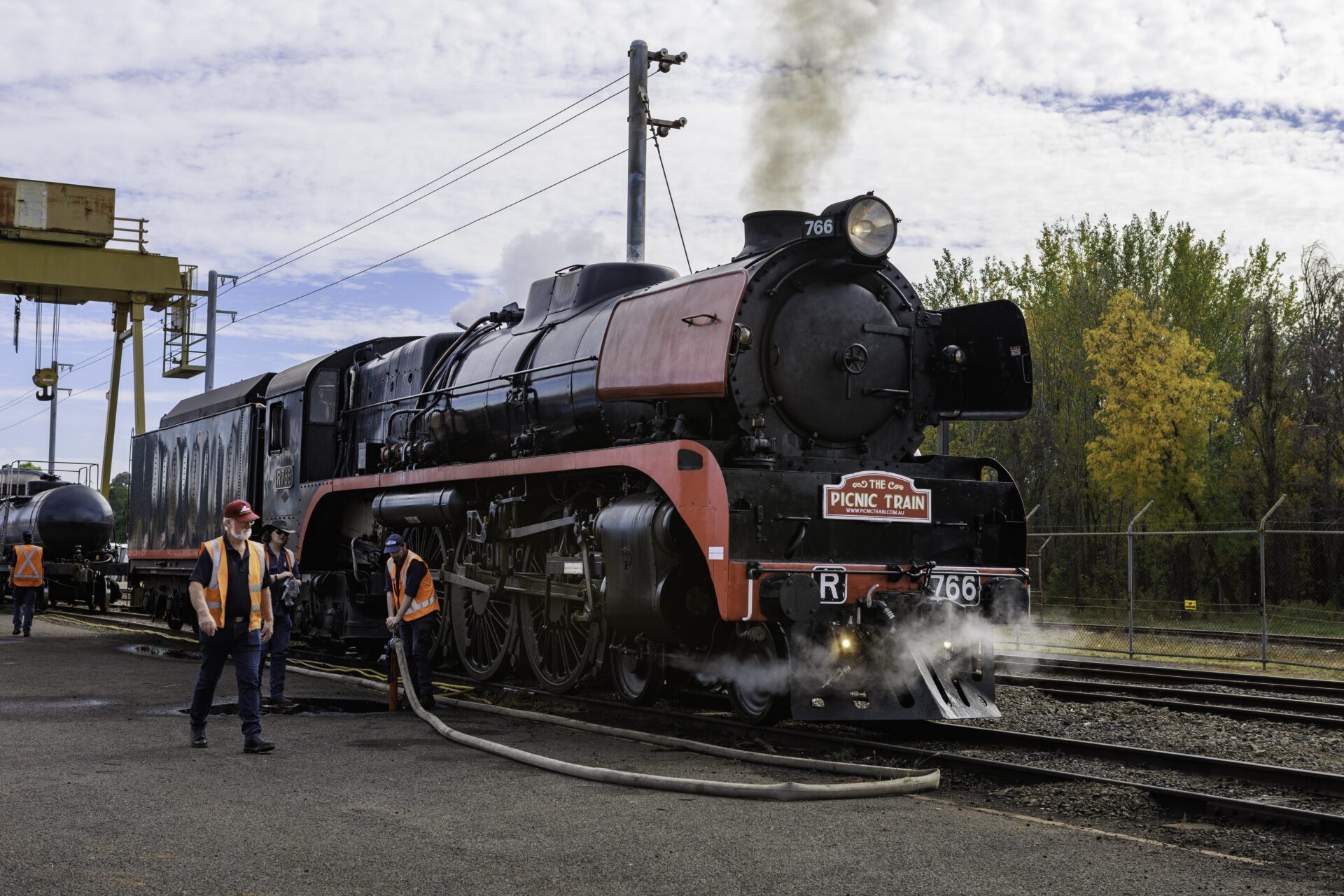 Steam locomotive "Picnic Train" with workers handling a hose beside it at a station.