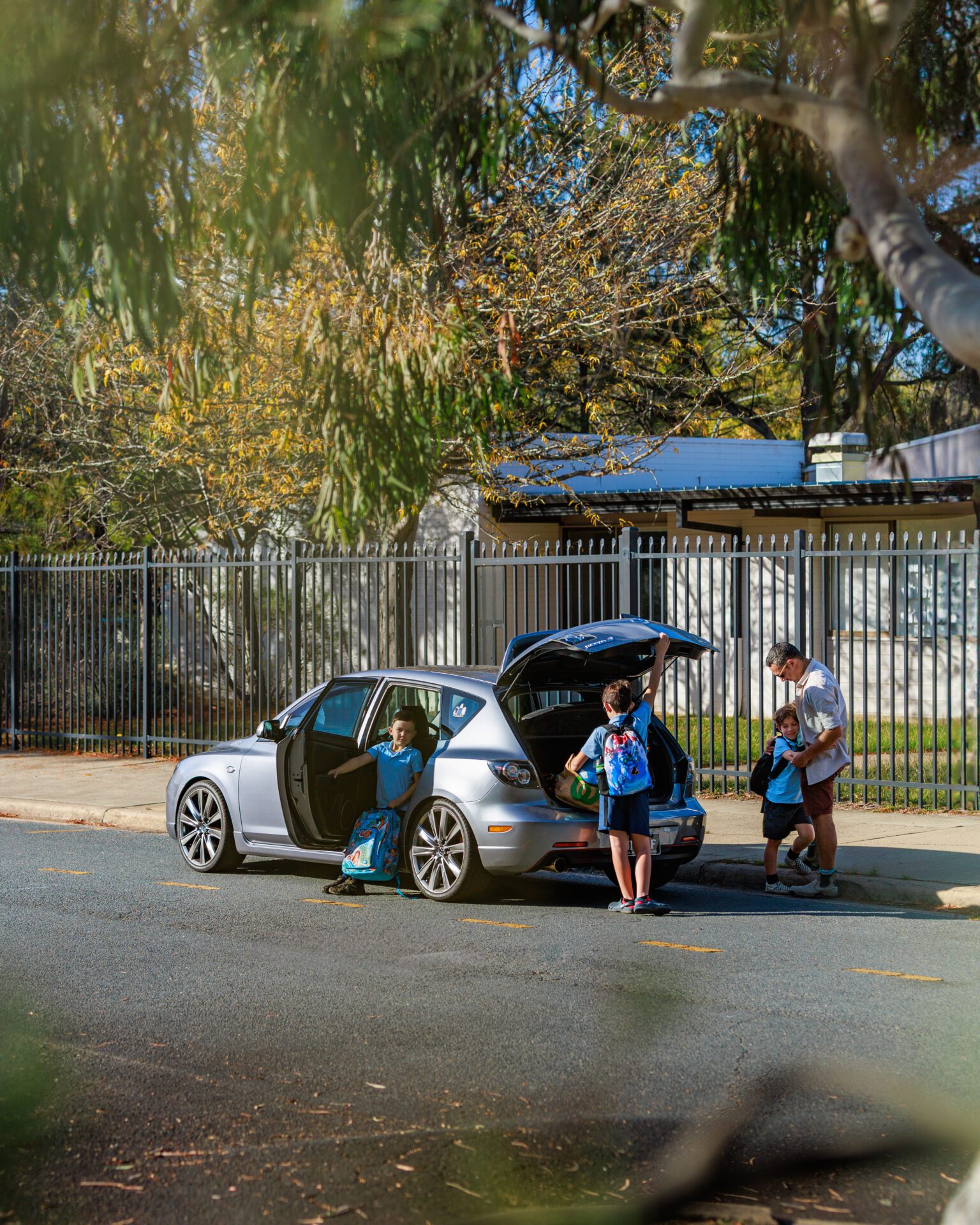 Father helps child by a silver car while two children with backpacks stand nearby on a sunny street.