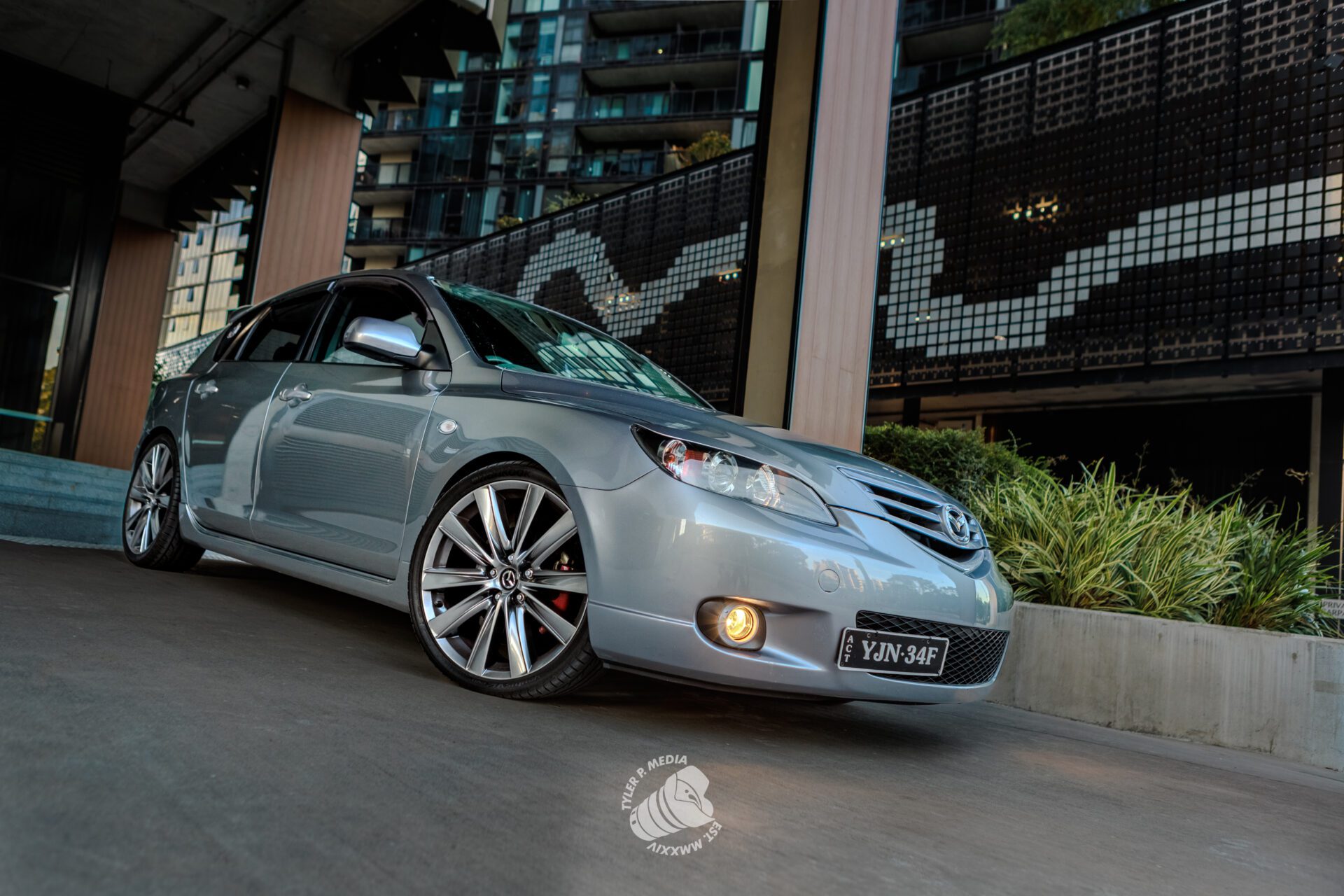 Silver Mazda sedan parked on a city street with modern buildings and greenery in background.