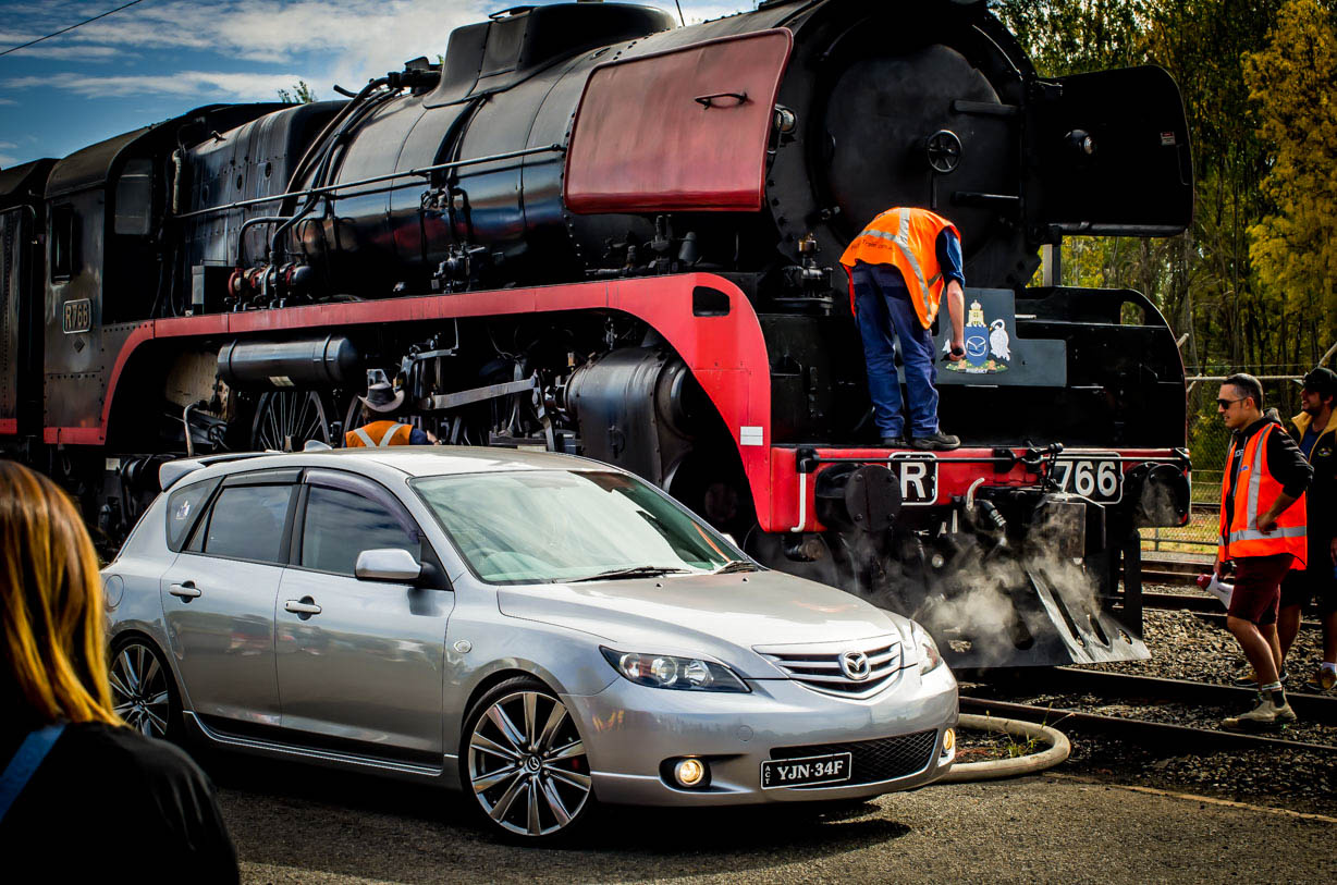 Silver Mazda car parked in front of a black and red steam locomotive with workers nearby.