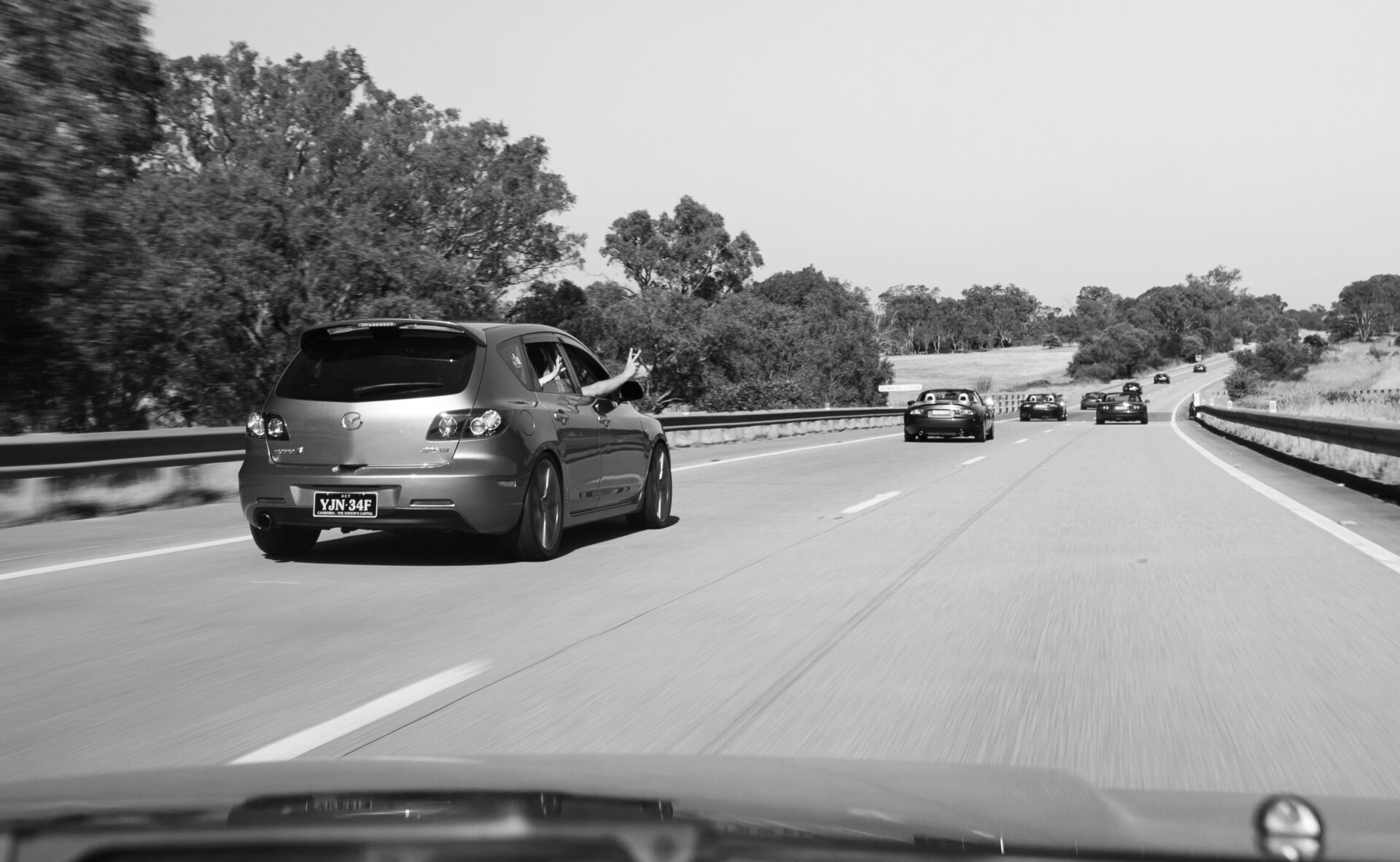 Black and white image of a highway with several cars driving, one car showing a hand gesture out the window.