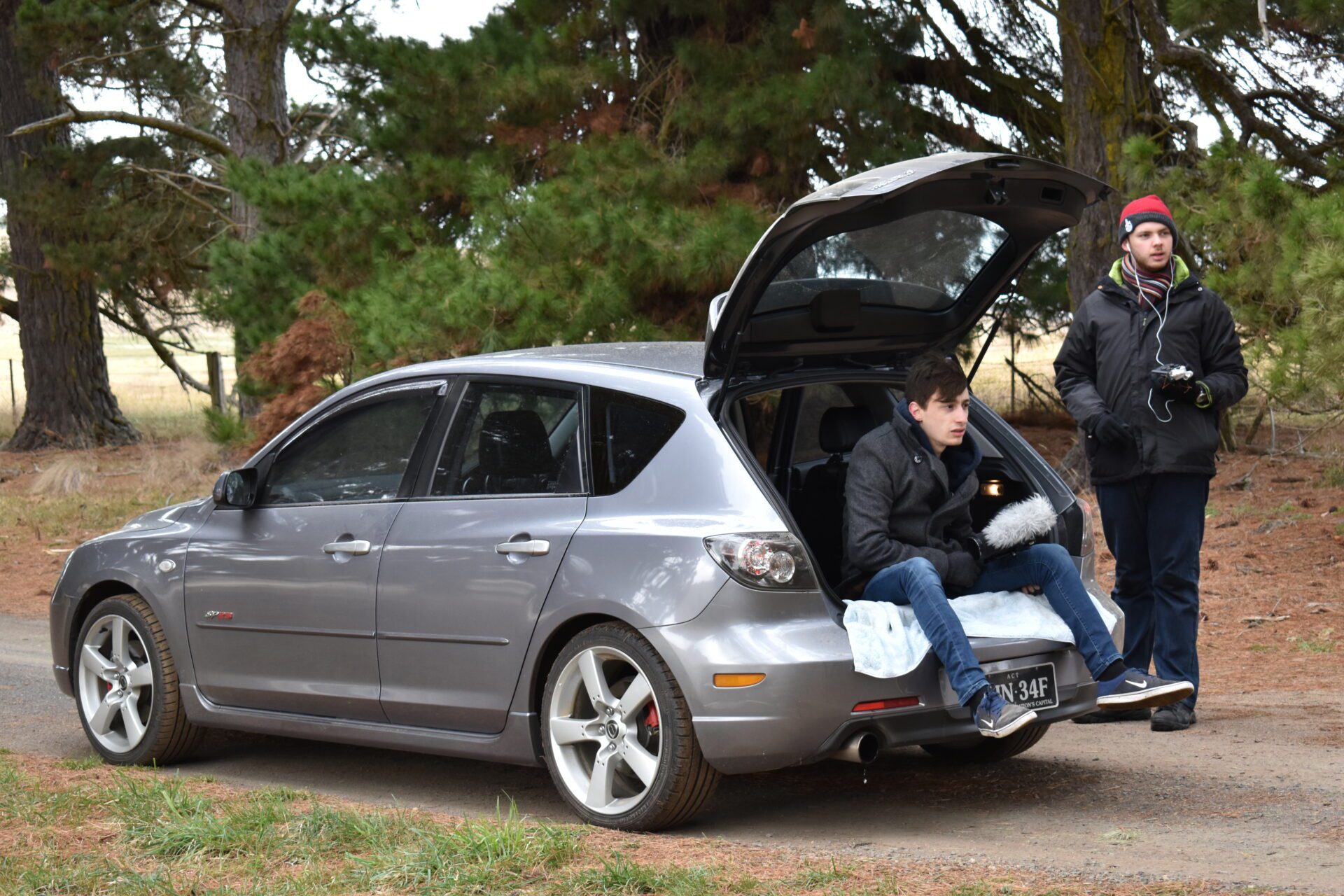 Two men with winter clothes near a gray hatchback car, one sitting in the open trunk holding a microphone.