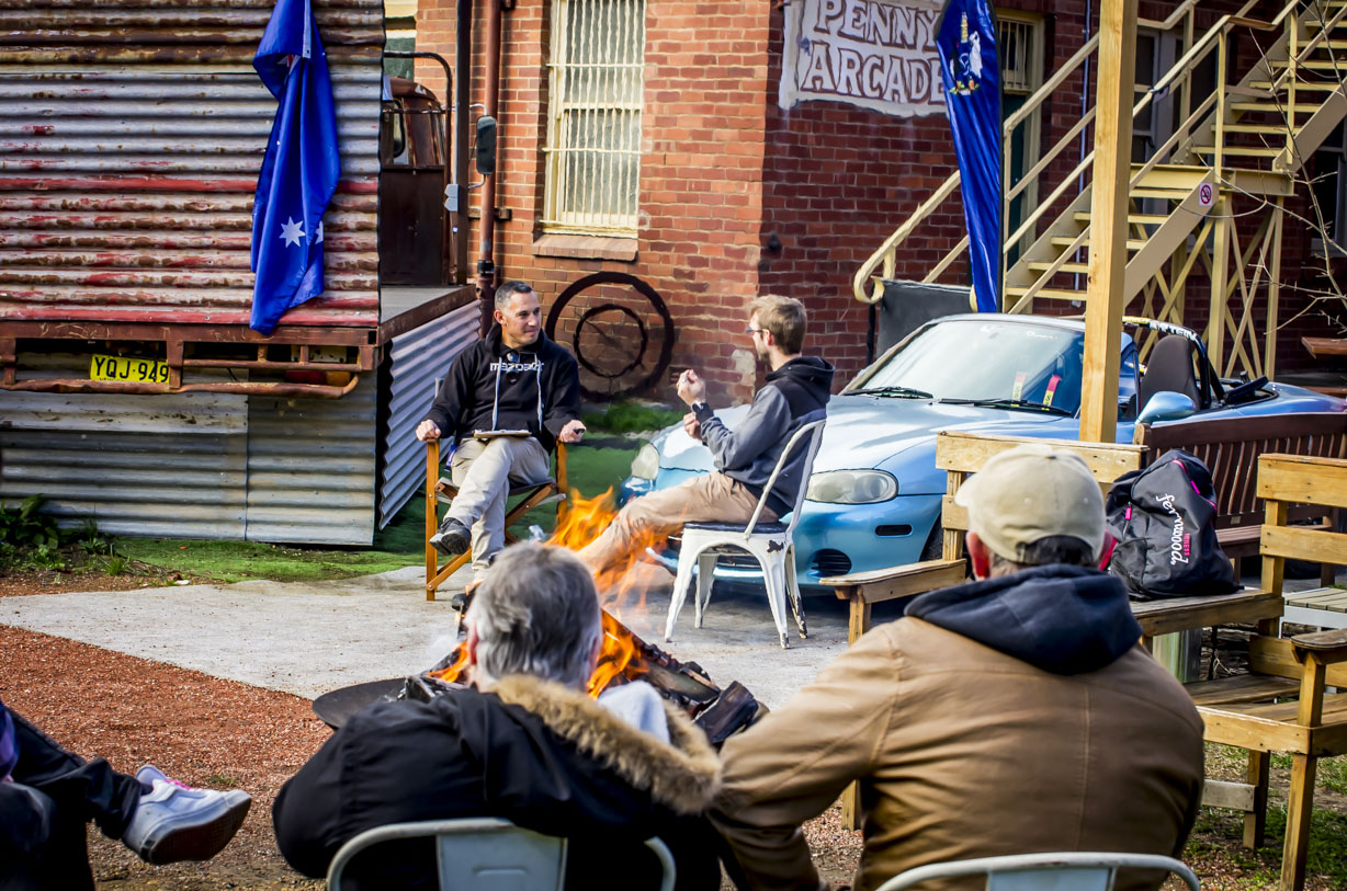 Group of people sitting around outdoor fire pit near blue car and brick building with "Penny Arcade" sign.