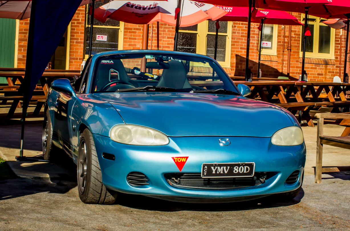 Blue Mazda convertible parked outdoors near picnic tables and umbrellas.