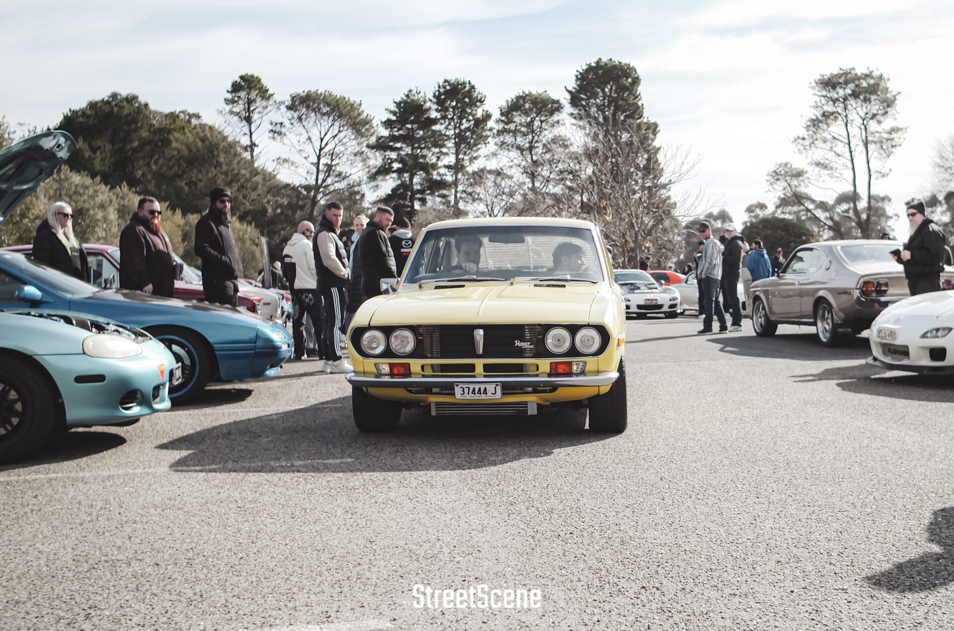 Yellow classic car in the center of a car show with people and other cars around.